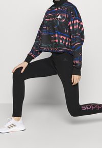 Osoba v černých legínách s růžovým logem Adidas, barevné bundě s černým vzorem a bílých teniskách Adidas, pózující ve výpadu.