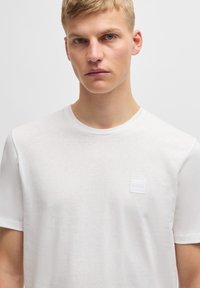 Witte t-shirt met korte mouwen van zacht stof, met een vierkant merkembleem in lichtgrijs op de linkerborst. Eenvoudig ontwerp, ronde hals.