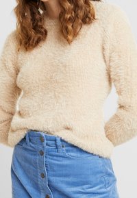 Beige fluffig stickad tröja med rund halsringning, kombinerad med en blå kordflorskjol med knappar framtill. Texturerad och mjuk.