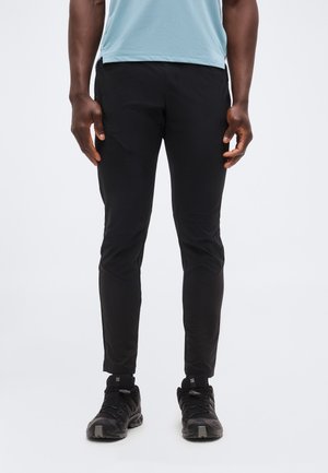 Férfi, fekete slim fit sportnadrágban és fekete tornacipőben, áll egy egyszerű világos háttér előtt.