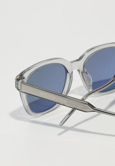 Ochelari de soare din plastic transparent cu lentile albastre, având un braț templu din metal argintiu cu marcaj gravat și accente texturate.