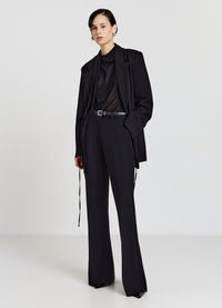 Blazer noir ajusté porté sur un top noir transparent et un pantalon noir taille haute. La tenue est équipée d'une ceinture et de jambes de pantalon évasées. Texture de tissu lisse.