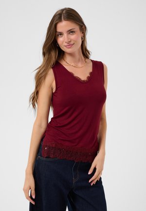 Kvinde med langt brunt hår iført en ærmeløs mørkerød top med blonder og mørkeblå jeans mod en ensfarvet lys baggrund.