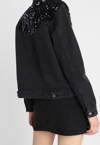 Mustat farkkutakki, jossa on paljettiyksityiskohtia hartioissa. Takissa on rento leikkaus ja nappisuljenta vyötäröllä, ja se on yhdistetty samasta kankaasta tehtyyn mustaan minihelmaan.