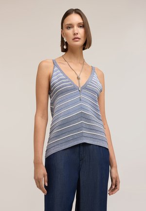 Femme portant un haut sans manches rayé bleu et blanc avec un col en V, un pantalon bleu foncé et un collier en argent sur un fond uni.