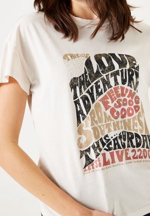 Femme portant un t-shirt blanc avec un texte au style vintage indiquant "The Love Adventure," "Feeling So Good," et les détails de l'événement en lettres noires, marron et rouges.