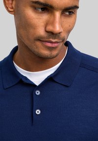 Granatowa koszulka polo wykonana z teksturowanej tkaniny, z klasycznym kołnierzykiem, plisą na dwa guziki z jasno niebieskimi guzikami, noszona na białym t-shircie.