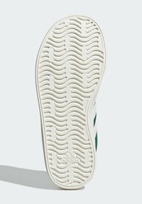 Suela de zapatilla blanca con patrón de tracción ondulado, que presenta una textura suave y un logo en relieve. Acentuada con rayas verdes a lo largo de los costados.