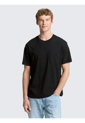 Jonge man met krullend haar die een zwart T-shirt met korte mouwen en een borstzak draagt en een lichtblauwe spijkerbroek, staand met één hand in de zak.