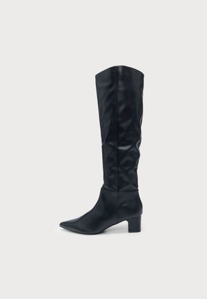 Botte noire en cuir montant jusqu’au genou, à bout pointu, talon bloc bas et tige droite, montrée de profil sur un fond clair.