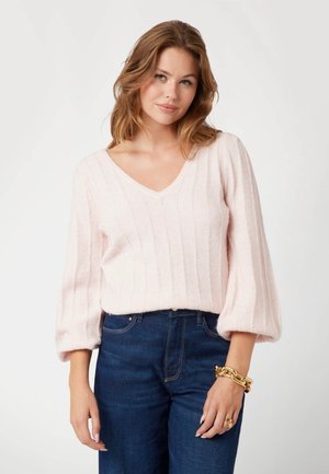 Maglione rosa chiaro lavorato a maglia con scollatura a V, texture a coste, e maniche lunghe a sbuffo, abbinato a jeans blu scuro a vita alta. Dettaglio con bracciale dorato.