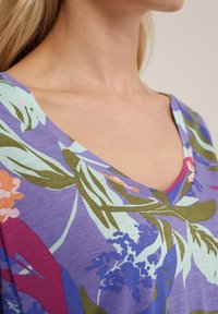 Top lyhythihaisena V-pääntiellä violetissa, jossa on kukkakuosi; sisältää vihreitä lehtiä, vaaleanpunaisia ja sinisiä kukkia sekä pehmeän, kevyen kankaan.