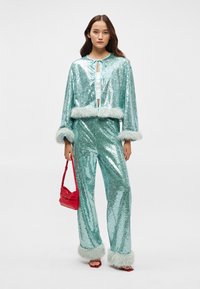 Conjunto em turquesa com lantejoulas e detalhes de pele sintética nas mangas e na bainha. O par inclui um casaco cropped e calças de perna larga. A mala vermelha adiciona um contraste.