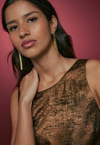 Ærmeløs, bronze kjole med en tekstureret overflade. Guld, lange hængende øreringe. Baggrunden er blød pink. Neutral makeup og naturlig hår.