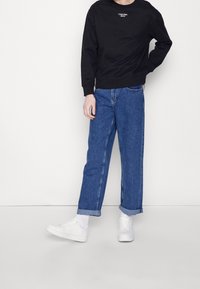 Persona que lleva puestas unas jeans azules con dobladillos, un sudadera negra de Calvin Klein, calcetines blancos y zapatillas blancas frente a un fondo blanco liso.
