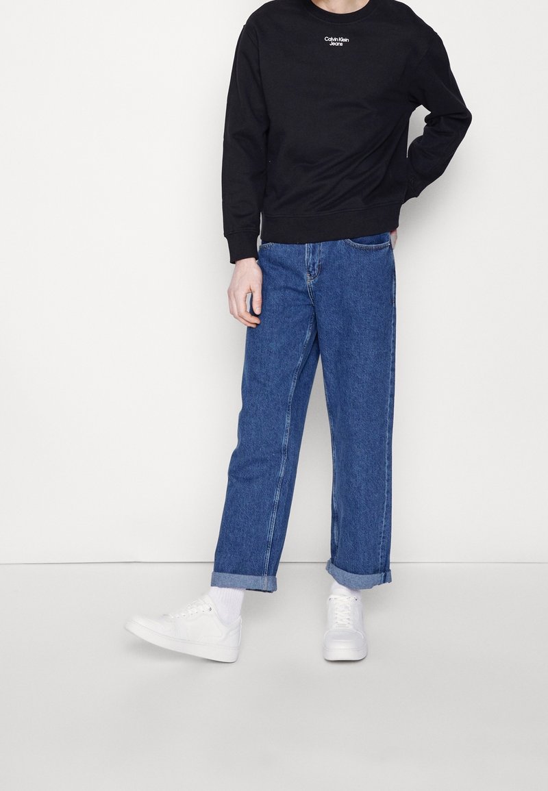 Persona que lleva puestas unas jeans azules con dobladillos, un sudadera negra de Calvin Klein, calcetines blancos y zapatillas blancas frente a un fondo blanco liso.