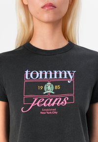 Černé bavlněné tričko s mnohobarevným grafickým textem: "tommy" v modré a růžové barvě, "jeans" v růžové barvě, s nápisem "1985" a logem vavřínu.