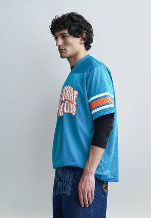 Ung mand med krøllet hår iført en blå sportstrøje over en sort langærmet skjorte og blå jeans, vendt mod venstre i en neutral omgivelser.