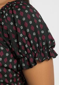 Svart tyg med röd och vit blommönster på en samlad puffärm som täcker en överarm mot en enfärgad ljus bakgrund.