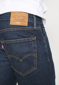 Tummansiniset farkut, joissa on nahkatarra, jossa lukee "Levi Strauss & Co." takavanteella, keltaiset tikkaukset ja muotoiltu takatasku.