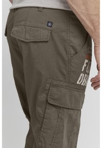 Khaki cargo-bukser med flere lommer, et struktureret stof og et mærke på bagsiden nær taljen. Har et trykt design på siden.