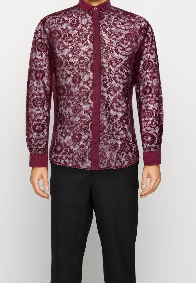 Homme portant une chemise en dentelle florale bordeaux à manches longues avec un col et des poignets unis, assortie à un pantalon noir.