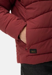 Czerwona ocieplana kurtka z pikowanym wzorem, wyposażona w zapinaną na zamek kieszeń i metkę z napisem "camel active, stworzona na codzienne przygody".