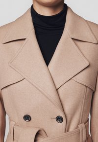 Beżowy płaszcz z włókien wełnianych z podwójnym zapięciem z przodu, dużymi klapami i dwoma czarnymi guzikami. Posiada pasek w talii i miękką teksturę.