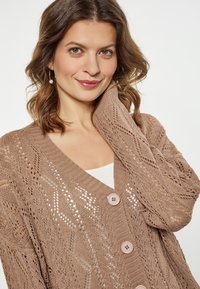 Lichtbruine gebreide cardigan met een geometrisch kantenpatroon, V-hals en vier ronde knopen aan de voorkant. Zachte textuur en ontspannen pasvorm.