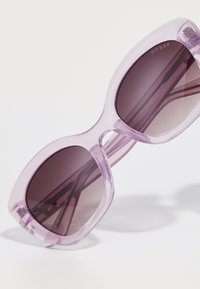 Gafas de sol moradas translúcidas con montura de gran tamaño, lentes oscuros con degradado y acentos de metal en las varillas. Logo de la marca visible en los lentes.