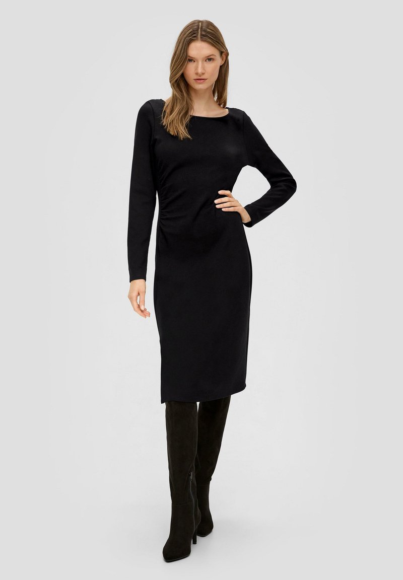Schwarzes langärmeliges Kleid mit taillierter Silhouette, ausgestattet mit einem Rundhalsausschnitt und Raffung am Taillenbereich. Kombiniert mit schwarzen knielangen Stiefeln.