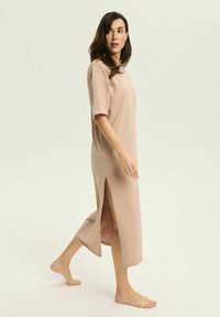 Jasno beżowa, dzianinowa sukienka do kolan, wykonana z miękkiego materiału. Posiada krótkie rękawy, detale w postaci rozcięcia z boku oraz luźny krój.