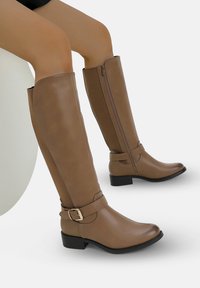 Botas de cuero marrón hasta la rodilla con cremallera lateral, detalle de correa con hebilla y tacón negro bajo, en una persona sentada con las piernas cruzadas.