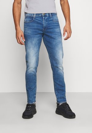 Mann trägt eng anliegende blaue Jeans mit ausgewaschenen Details, weiße Socken, schwarze Sneakers und ein hellgraues T-Shirt vor einfachem Hintergrund.