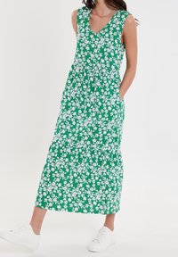 Robe maxi verte à motifs floraux blancs, sans manches, décolletée en v, taille froncée et poches ; associée à des baskets blanches.