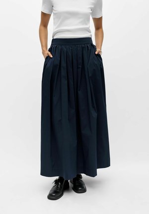 Femme portant un haut blanc à manches courtes, une longue jupe plissée foncée avec poches et des chaussures noires à lacets sur un fond uni.
