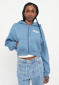 Moletom curto azul feito de tecido macio, com fecho de correr na frente, grandes bolsos e bordado em texto branco no peito.