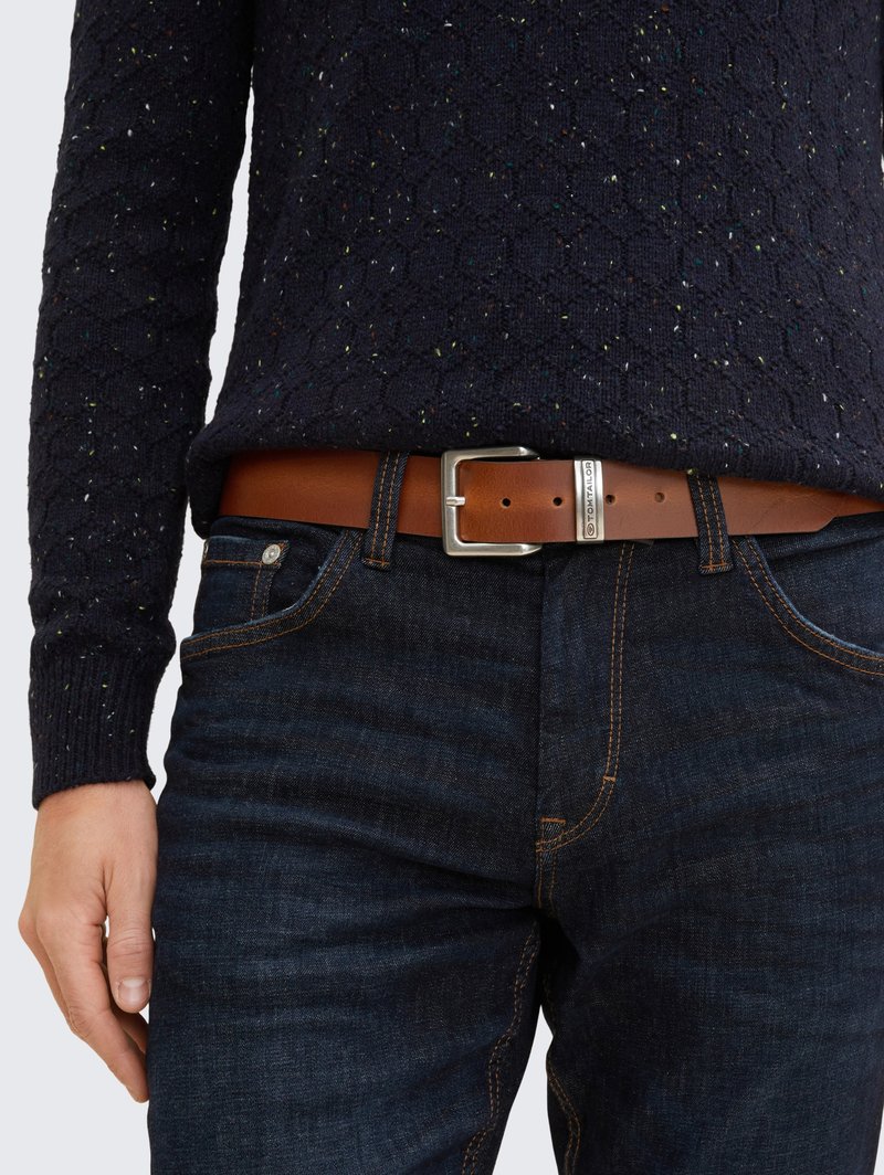 Brauner Ledergürtel mit silberner rechteckiger Schnalle, getragen über dunklen Denim-Jeans, kombiniert mit einem marineblauen Strickpullover mit strukturiertem Muster.