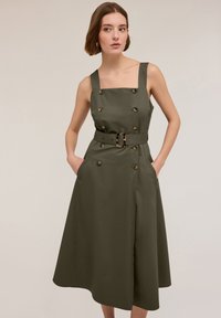 Olivgrön klänning med fyrkantig halsringning, breda axelband, dubbelknäppt front, midjebälte, två sidofickor och A-linjeformad kjol.
