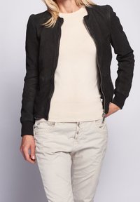 Svart perforerat läderjacka med figurnära design, ribbade ärmslut och zip-stängning. Kombineras med en krämfärgad tröja och beiga byxor.