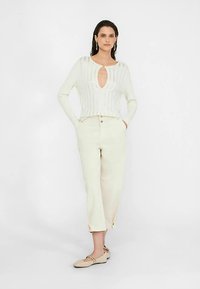 Biely rebrovaný pletený sveter s výstrihom na kľúčik, v kombinácii s vysokými krémovými nohavicami, majúcimi rovný strih a rázporky pri členkoch.