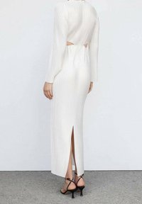 Robe midi blanche à côtes avec manches longues, découpe dans le dos et fente sur le côté. Associée à des sandales à talons noires à lanières. Texture lisse, silhouette ajustée.