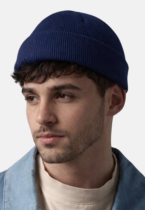 Junger Mann mit kurzen dunklen Haaren und leichtem Bart, trägt eine marineblaue, gerippte Mütze, ein beiges Hemd und eine hellblaue Jacke, schaut leicht nach links.