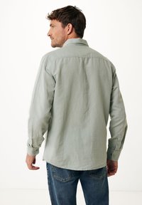 Světle zelená košile na knoflíky s uvolněným střihem, zadním dílem a dlouhými rukávy, v kombinaci s modrými džíny, na pozadí bílé plochy.