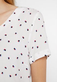 Top blanco de manga corta con escote en V, tejido texturizado y un patrón de pequeñas formas geométricas en rojo, azul y negro.