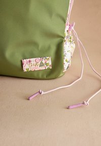 Rucsac verde cu accente florale, având un etichetă, sfori roz și o textură netedă. Baza are un design vibrant, cu modele.