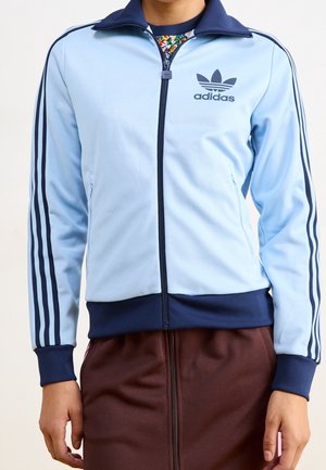 Jasnoniebieska kurtka Adidas zapinana na zamek błyskawiczny z granatowymi paskami na rękawach i granatowym kołnierzem, noszona z brązową spódnicą.