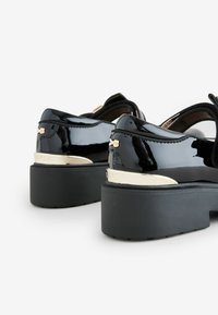 Zwarte gepatenteerde leren plateauschoenen met gouden accenten en een dikke zwarte zool. Voorzien van een ronde teen en een uitsnijding aan de achterkant.