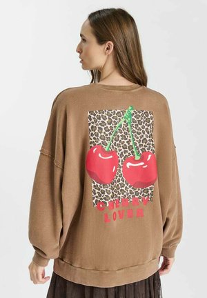 Vrouw met lang bruin haar draagt een bruin oversized trui met luipaardprint en twee rode kersen boven de tekst "Cherry Lover" op de achterkant.