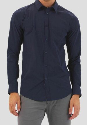 Homme portant une chemise bleu foncé ajustée à manches longues boutonnée avec un pantalon gris, debout devant un fond clair uni.